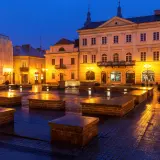 Piotrków Trybunalski atrakcje: Rynek nocą, mokry bruk odbija światła latarni i budynków. Kamienne ławki tworzą ciekawe formy.