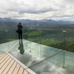 Mężczyzna na szklanej platformie wieży widokowej podziwia panoramę gór.