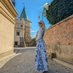Kobieta w sukience w kwiaty podziwia biecz atrakcje. W tle zamek i brukowana uliczka.