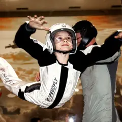 Chłopiec w kasku i kombinezonie doświadcza lotów w tunelu aerodynamicznym, unosząc się w powietrzu pod okiem instruktora.