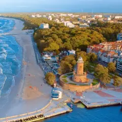 Widok z lotu ptaka na promenadę nad morzem z piaszczystą plażą, falującym morzem, latarnią morską i portem z łodziami.