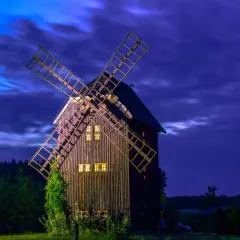 Drewniany wiatrak nocą, oświetlony ciepłym światłem z okien, wśród zieleni. To jedno z tych czarodziejskich miejsc w Polsce, które budzą wyobraźnię.