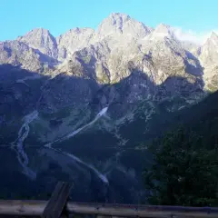 Morskie Oko odbija się w spokojnej tafli wody. Majestatyczne góry, oświetlone słońcem, wznoszą się na tle błękitnego nieba. Wysokość nad poziomem morza robi wrażenie.
