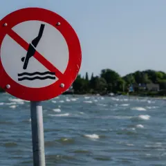 Znak zakazu kąpieli nad morzem. Dwie osoby w wodzie ignorują ostrzeżenie.