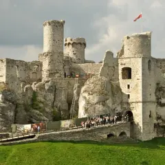 Imponujące ruiny zamku na skale, z polską flagą na wieży. Tłum turystów zwiedza zamek.