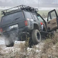 Czarny Jeep z bagażnikiem dachowym pokonuje trudny teren, to prawdziwe off road warszawa! Dym z opon i otwarta drzwi dodają dynamiki.