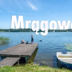 Mrągowo atrakcje: spokojny dzień nad jeziorem, dzieci łowią ryby z pomostu, żaglówki na horyzoncie.