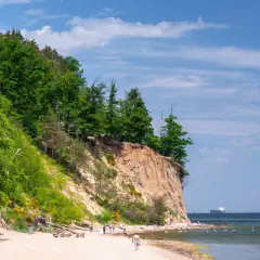 Piękny dzień nad polskim morzem. Klif porośnięty drzewami, piaszczysta plaża z kilkoma spacerującymi ludźmi i statkiem na horyzoncie.