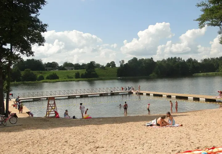 Słoneczne plaże na Mazurach tętnią życiem. Ludzie odpoczywają na piasku, kąpią się w jeziorze i spacerują po pomoście.