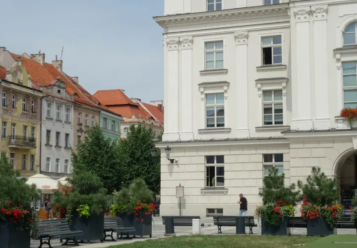 Kalisz atrakcje: biały budynek z kolumnami, kamienice z czerwonymi dachami, ławki i zielone donice z kwiatami.