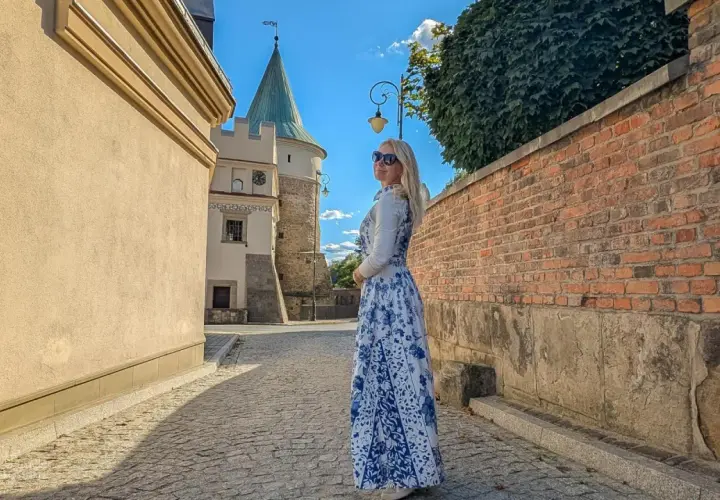 Kobieta w sukience w kwiaty podziwia biecz atrakcje. W tle zamek i brukowana uliczka.