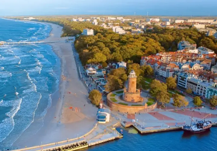 Widok z lotu ptaka na promenadę nad morzem z piaszczystą plażą, falującym morzem, latarnią morską i portem z łodziami.