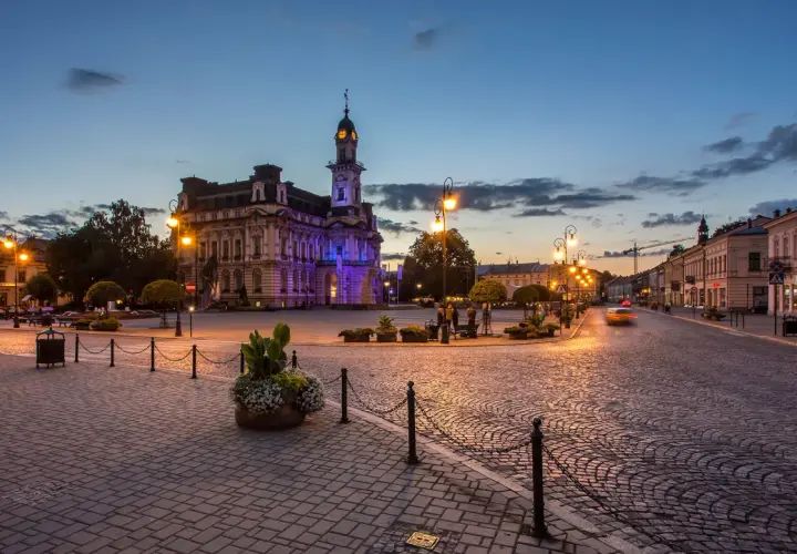 Wieczorny rynek w Nowym Sączu, oświetlony latarniami i iluminacją ratusza. Brukowana nawierzchnia i zabytkowa architektura tworzą niepowtarzalny klimat, zachęcając do odkrywania atrakcji Nowego Sącza.