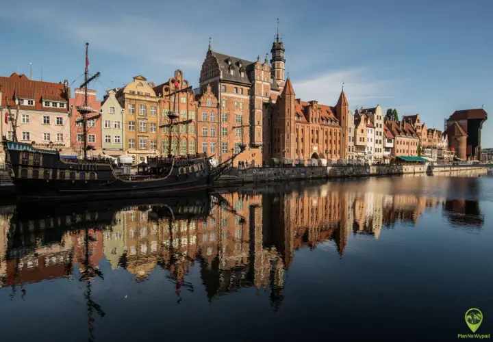 Gdańsk atrakcje: zabytkowy żaglowiec cumuje przy Motławie, w tle kolorowe kamienice i Żuraw. Odbicia budynków w wodzie.