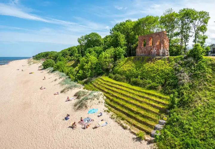 Piękna plaża z ruinami na klifie, zielone tarasy i ludzie odpoczywający na piasku. Trzęsacz atrakcje zachwycają!