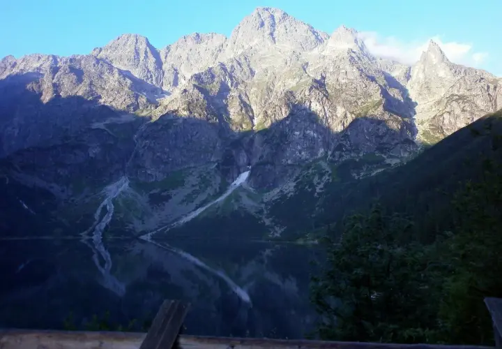Morskie Oko odbija się w spokojnej tafli wody. Majestatyczne góry, oświetlone słońcem, wznoszą się na tle błękitnego nieba. Wysokość nad poziomem morza robi wrażenie.