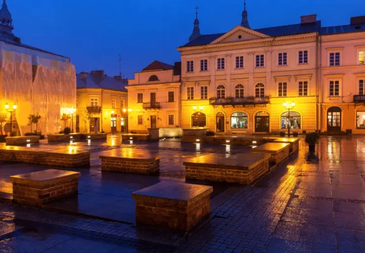 Piotrków Trybunalski atrakcje: Rynek nocą, mokry bruk odbija światła latarni i budynków. Kamienne ławki tworzą ciekawe formy.