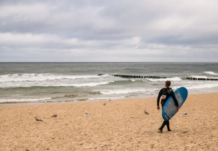 Surfer z deską na plaży w Juracie, szuka fal. W tle morze i mewy. Jedna z atrakcji nad polskim morzem.