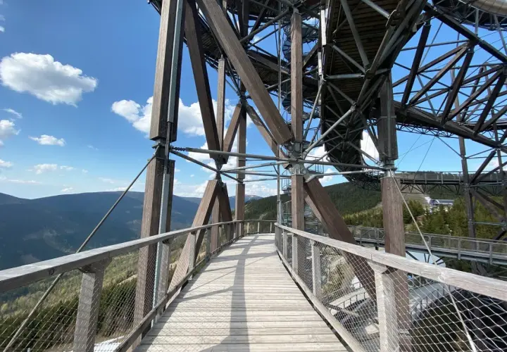 Drewniany sky bridge wznosi się nad górami, oferując widok na lasy i błękitne niebo z chmurami.