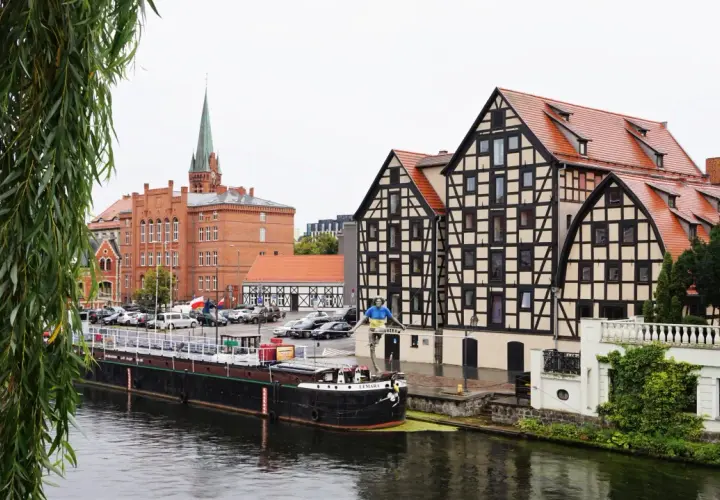 Bydgoszcz atrakcje: nad Brdą, w tle zabytkowe kamienice i kościół. Na pierwszym planie barka i rzeźba.