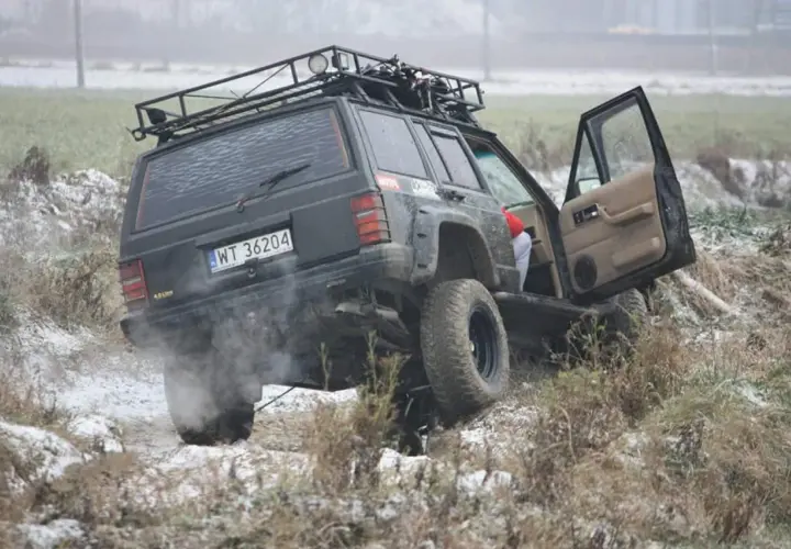 Czarny Jeep z bagażnikiem dachowym pokonuje trudny teren, to prawdziwe off road warszawa! Dym z opon i otwarta drzwi dodają dynamiki.