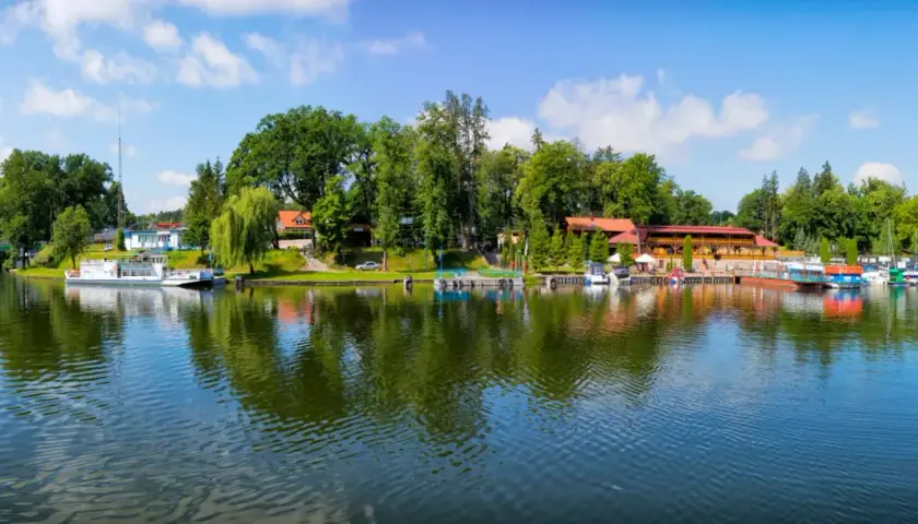 Malowniczy widok na port i jezioro w Rucianem Nidzie. Widać tu liczne łodzie, restauracje i zielone drzewa, tworzące idealne miejsce na wypoczynek i odkrywanie atrakcji.