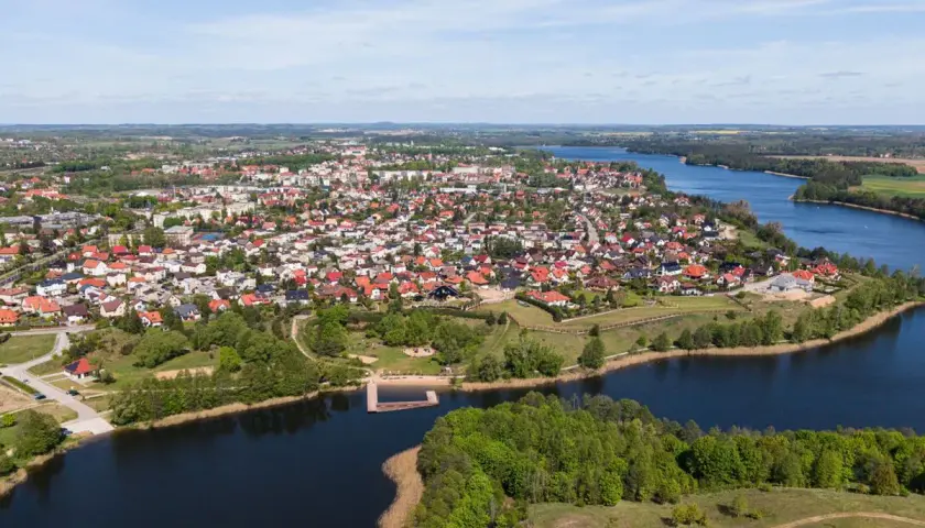 Widok z lotu ptaka na miasteczko nad jeziorem, jedno z największych rynków w Polsce, z domami z czerwonymi dachami i zielenią.