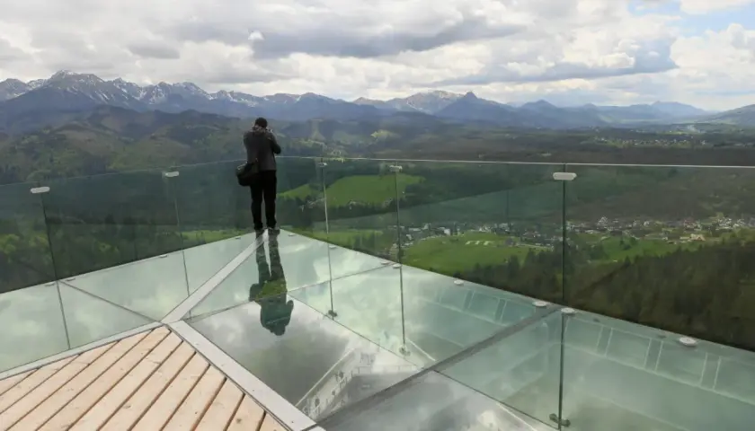 Mężczyzna na szklanej platformie wieży widokowej podziwia panoramę gór.