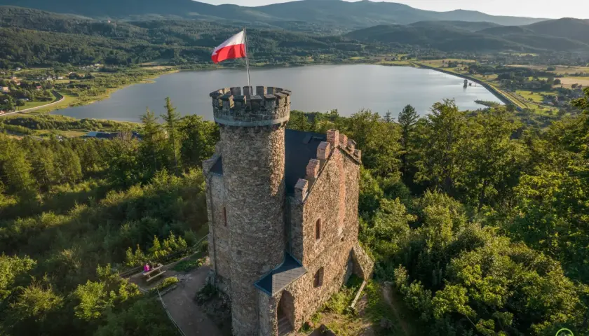 Kamienna wieża z polską flagą na szczycie, w tle jezioro i góry. Idealne miejsce, by odkryć, co warto zobaczyć w Sudetach.