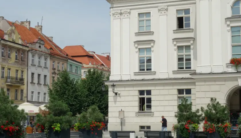 Kalisz atrakcje: biały budynek z kolumnami, kamienice z czerwonymi dachami, ławki i zielone donice z kwiatami.