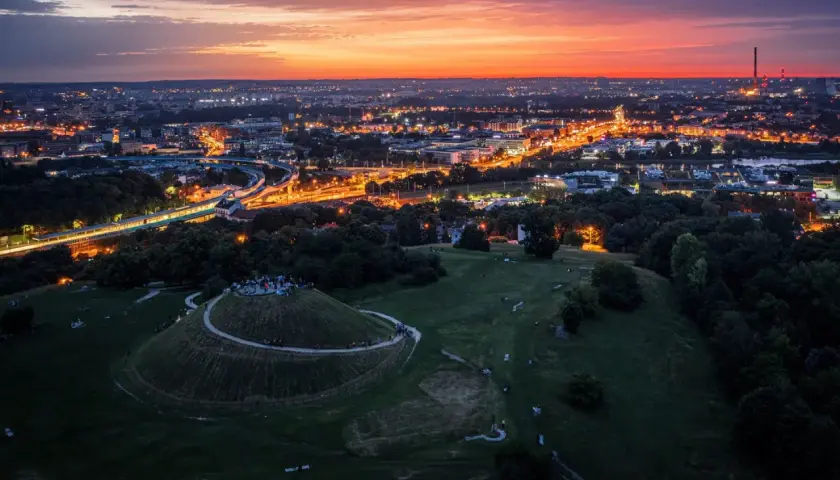 Zachód słońca nad Krakowem, z Kopcem Kraka w centrum uwagi. Miasto rozświetlone tysiącami świateł, a na wzgórzu gromadzą się ludzie.