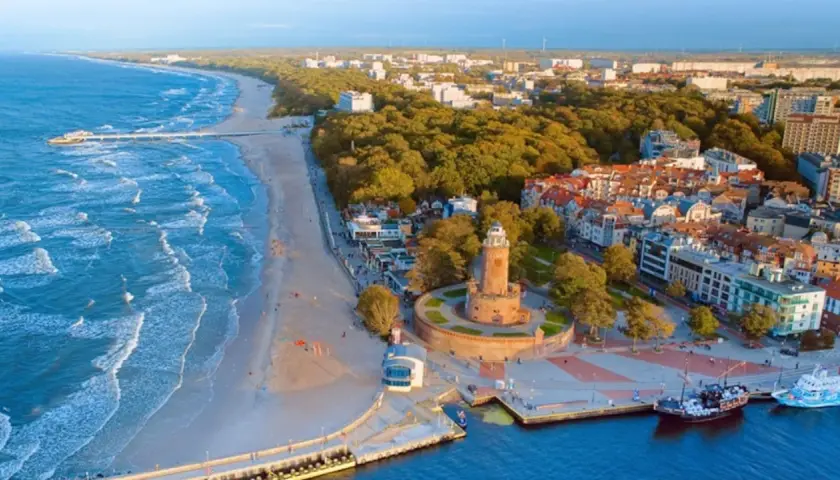 Widok z lotu ptaka na promenadę nad morzem z piaszczystą plażą, falującym morzem, latarnią morską i portem z łodziami.