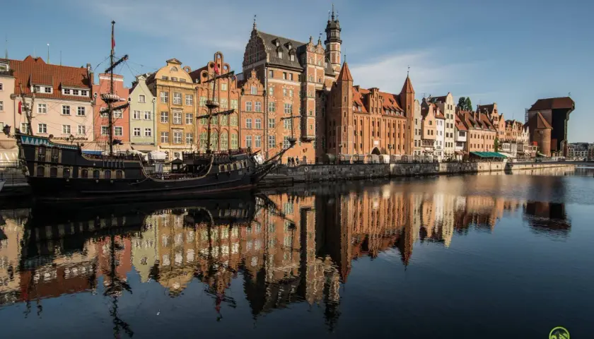 Gdańsk atrakcje: zabytkowy żaglowiec cumuje przy Motławie, w tle kolorowe kamienice i Żuraw. Odbicia budynków w wodzie.