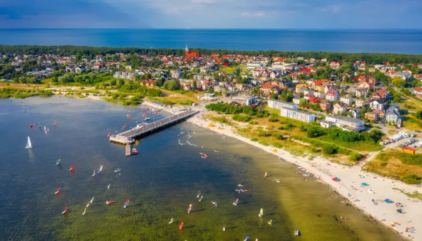 Jastarnia atrakcje: molo, plaża z turystami i liczne żaglówki oraz deski windsurfingowe na wodzie.