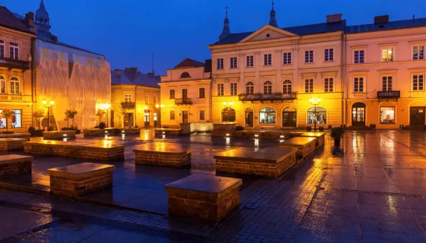 Piotrków Trybunalski atrakcje: Rynek nocą, mokry bruk odbija światła latarni i budynków. Kamienne ławki tworzą ciekawe formy.