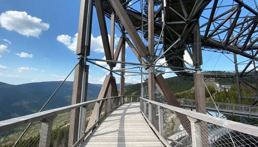 Drewniany sky bridge wznosi się nad górami, oferując widok na lasy i błękitne niebo z chmurami.