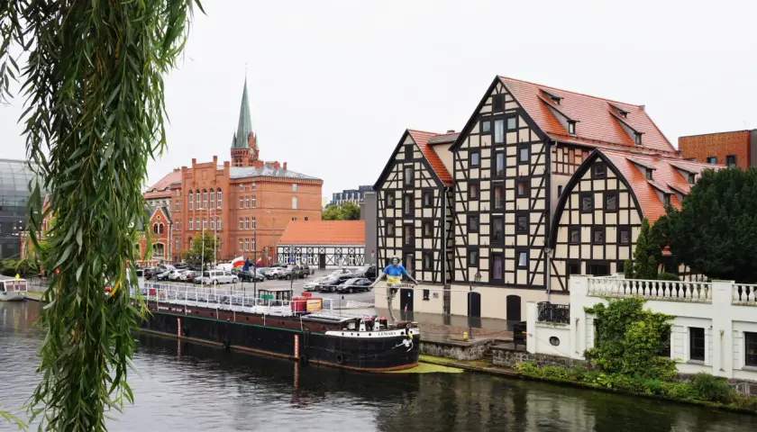 Bydgoszcz atrakcje: nad Brdą, w tle zabytkowe kamienice i kościół. Na pierwszym planie barka i rzeźba.