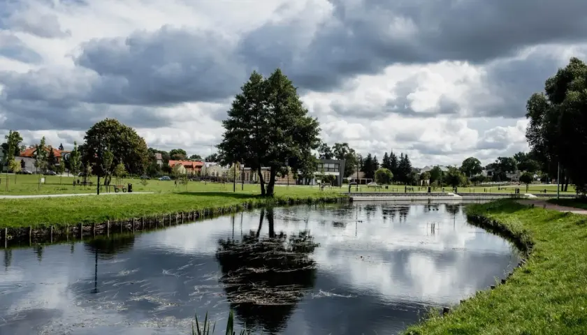 Spokojny park miejski w Suwałkach z jeziorem, drzewami i budynkami w tle. Idealne miejsce na relaks i odkrywanie suwalskich atrakcji.