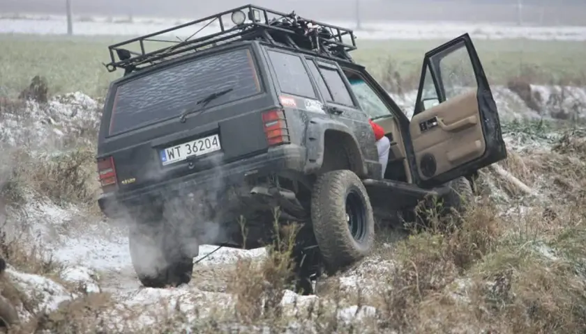 Czarny Jeep z bagażnikiem dachowym pokonuje trudny teren, to prawdziwe off road warszawa! Dym z opon i otwarta drzwi dodają dynamiki.
