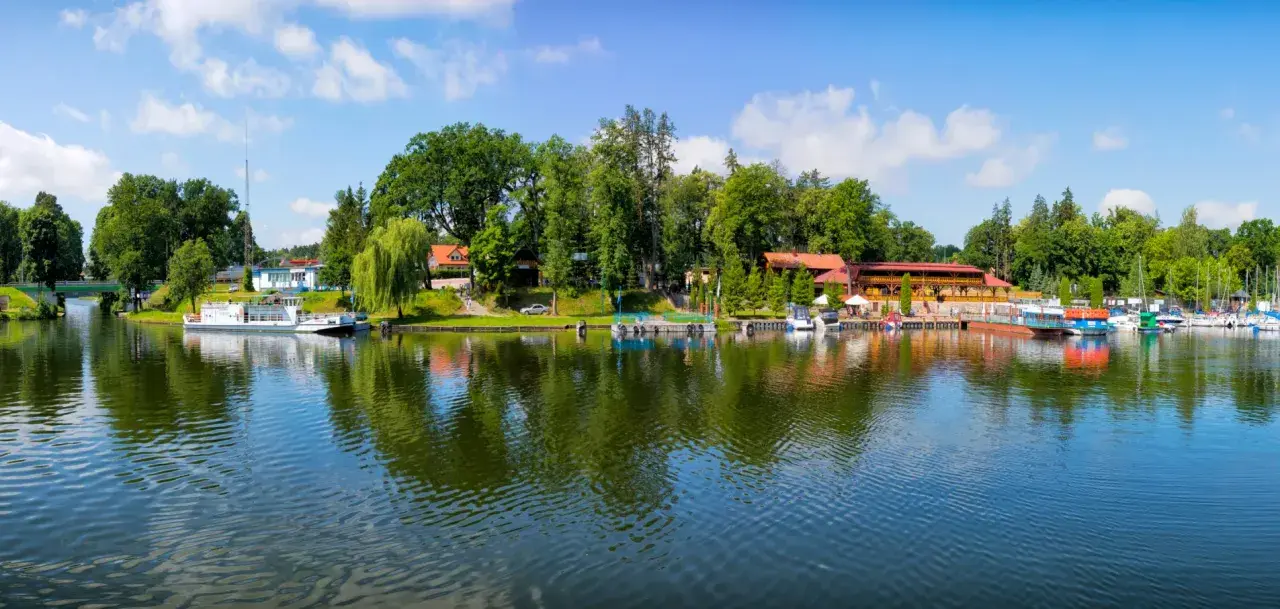 Malowniczy widok na port i jezioro w Rucianem Nidzie. Widać tu liczne łodzie, restauracje i zielone drzewa, tworzące idealne miejsce na wypoczynek i odkrywanie atrakcji.