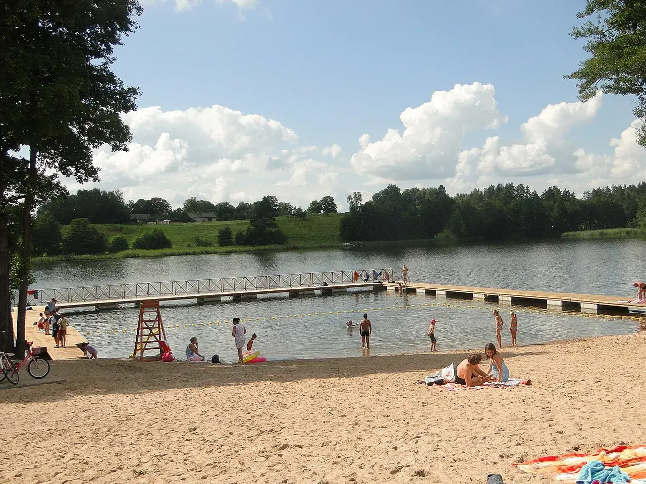 Słoneczne plaże na Mazurach tętnią życiem. Ludzie odpoczywają na piasku, kąpią się w jeziorze i spacerują po pomoście.