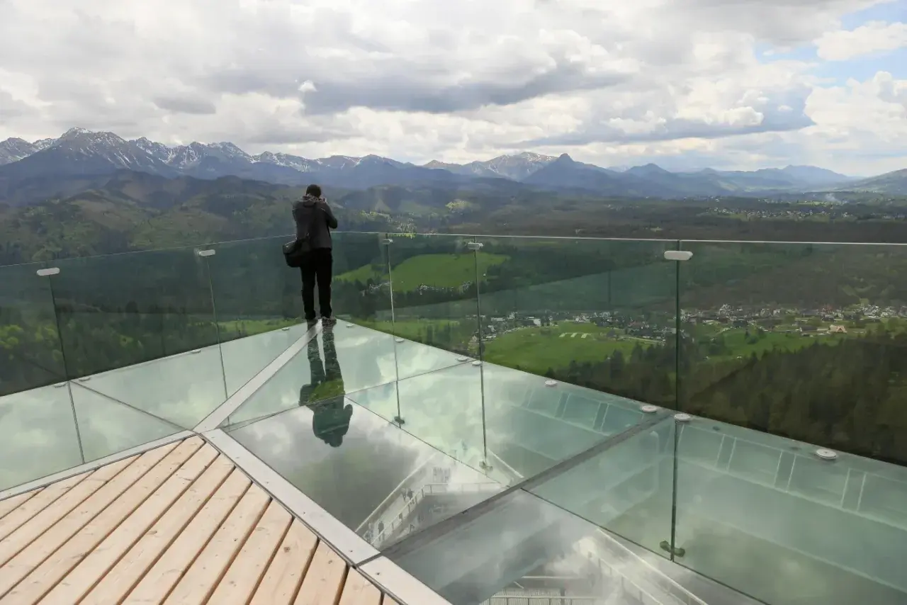 Mężczyzna na szklanej platformie wieży widokowej podziwia panoramę gór.