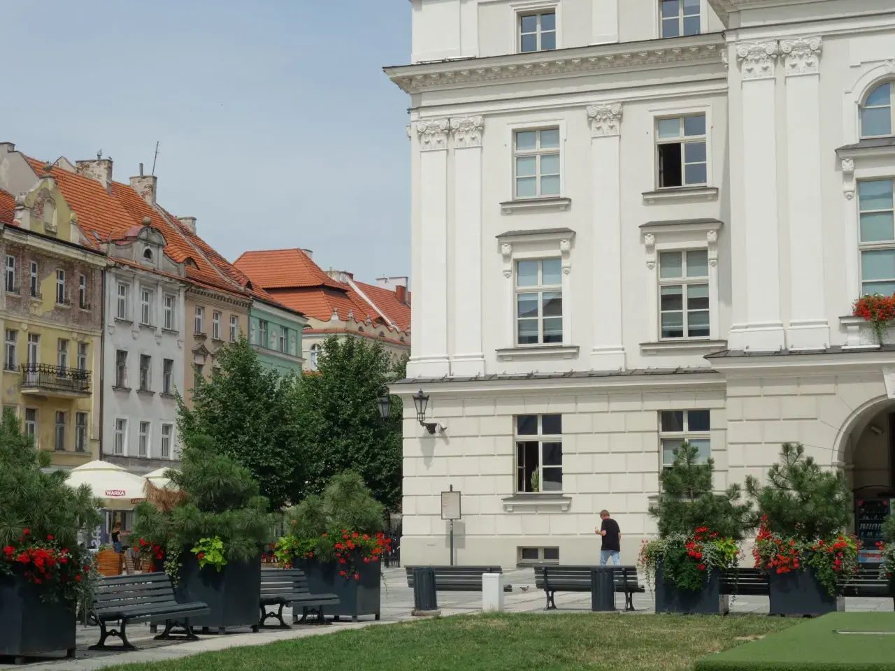 Kalisz atrakcje: biały budynek z kolumnami, kamienice z czerwonymi dachami, ławki i zielone donice z kwiatami.