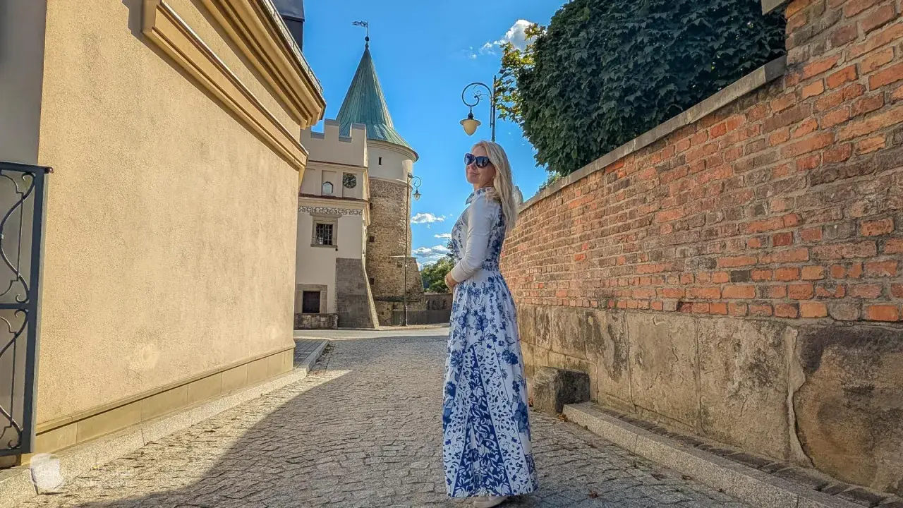 Kobieta w sukience w kwiaty podziwia biecz atrakcje. W tle zamek i brukowana uliczka.