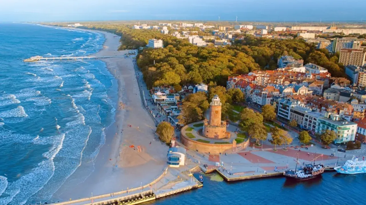 Widok z lotu ptaka na promenadę nad morzem z piaszczystą plażą, falującym morzem, latarnią morską i portem z łodziami.