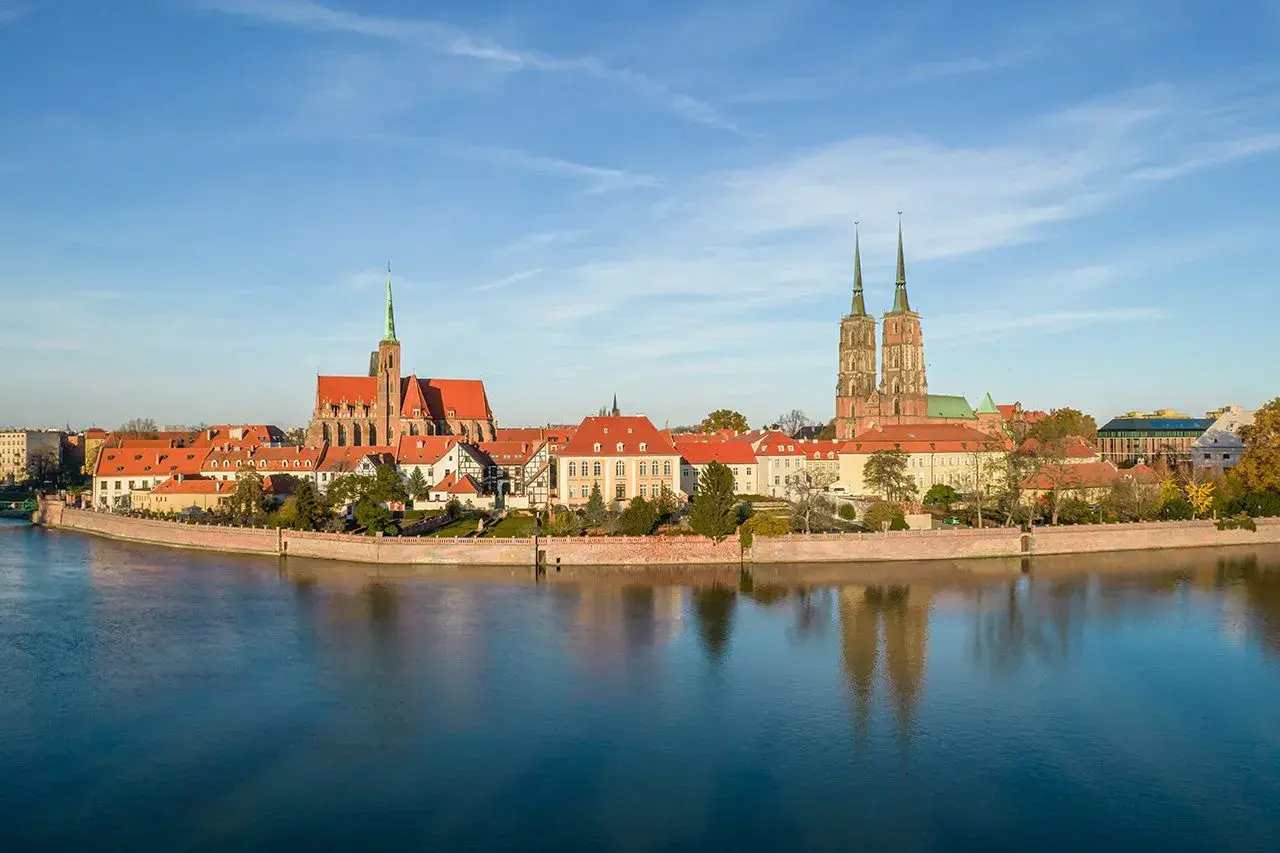 Widok na Ostrów Tumski, serce starego miasta Wrocławia, z katedrą i kościołem św. Krzyża odbijającymi się w spokojnej wodzie Odry.