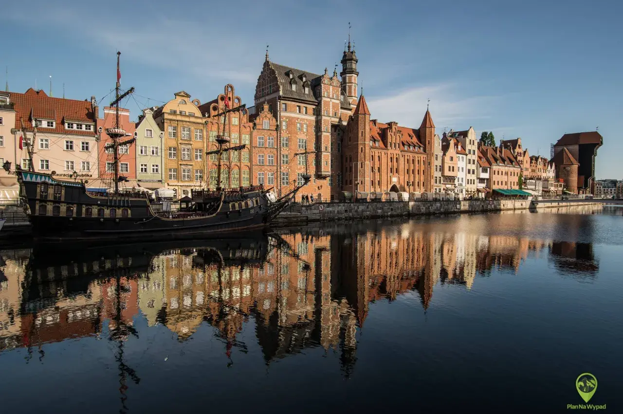 Gdańsk atrakcje: zabytkowy żaglowiec cumuje przy Motławie, w tle kolorowe kamienice i Żuraw. Odbicia budynków w wodzie.