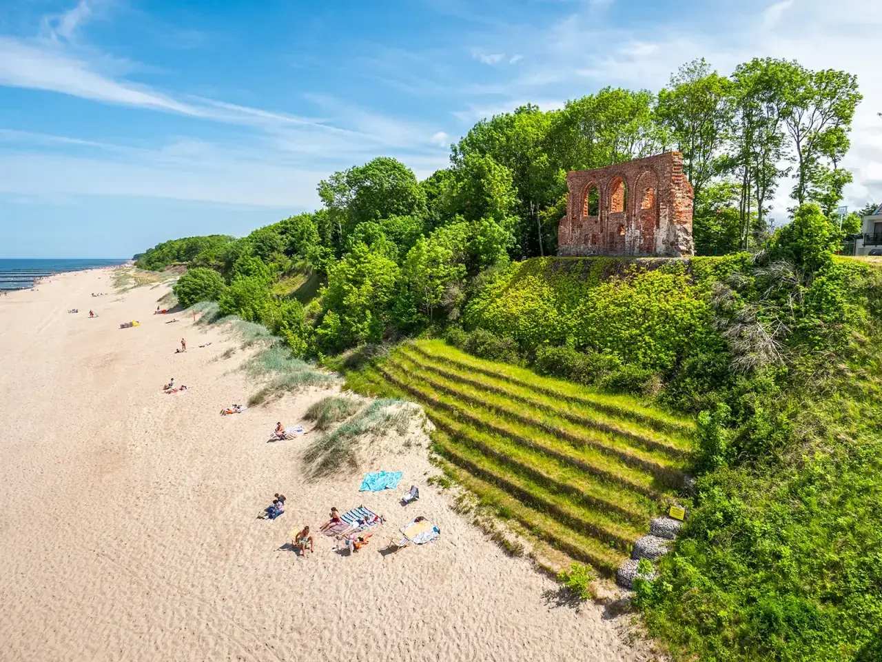 Piękna plaża z ruinami na klifie, zielone tarasy i ludzie odpoczywający na piasku. Trzęsacz atrakcje zachwycają!