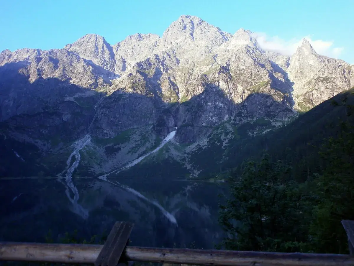 Morskie Oko odbija się w spokojnej tafli wody. Majestatyczne góry, oświetlone słońcem, wznoszą się na tle błękitnego nieba. Wysokość nad poziomem morza robi wrażenie.