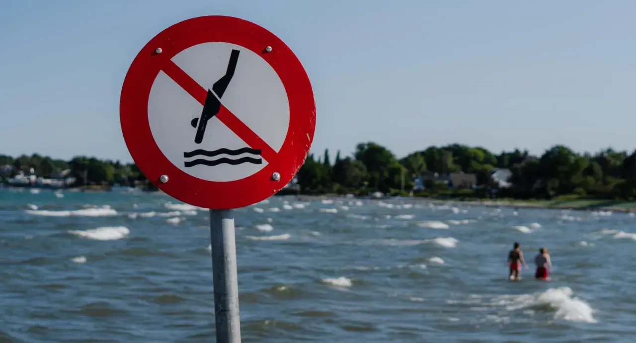 Znak zakazu kąpieli nad morzem. Dwie osoby w wodzie ignorują ostrzeżenie.